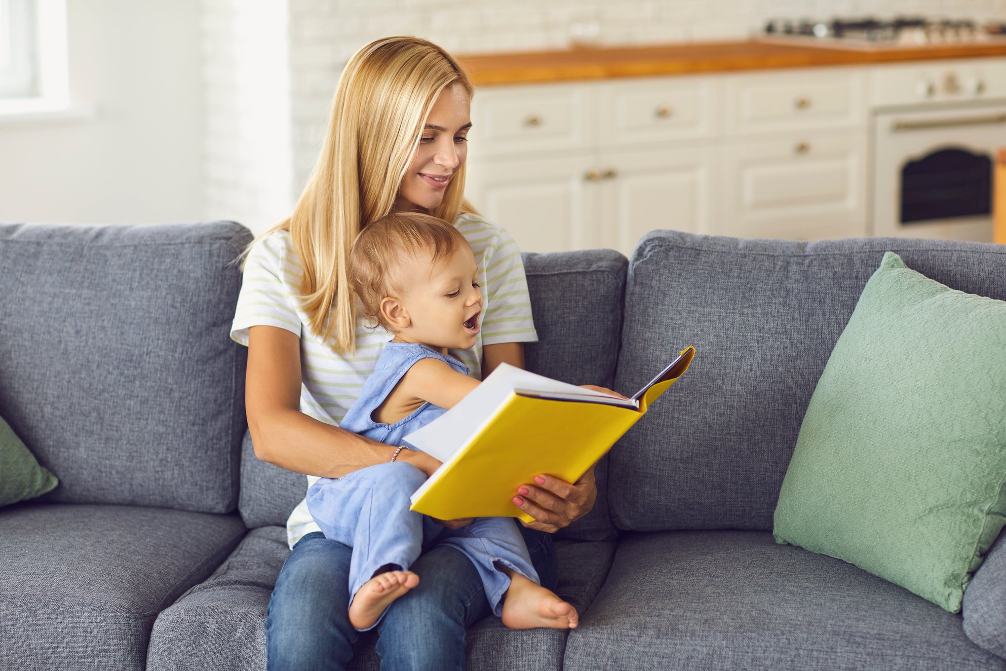 Young Mommy Reading Book of Fairy Tales to Her Little Child Sitting on Couch at Home